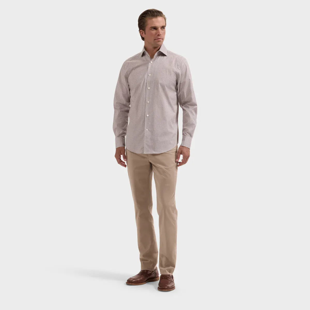 A man modeling a light beige long-sleeve button-down dress shirt with beige trousers and brown leather shoes, standing against a solid light gray background.