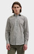 A man wearing a long-sleeve button-up shirt with a dense micro-pattern in black, white, and gray, paired with olive green trousers, shot against a solid light gray background.