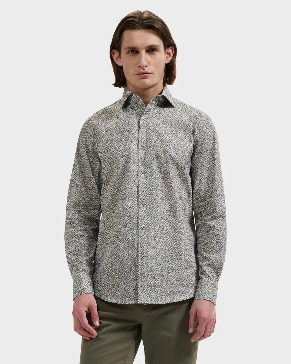 A man wearing a long-sleeve button-up shirt with a dense micro-pattern in black, white, and gray, paired with olive green trousers, shot against a solid light gray background.