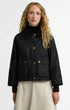 A model with long curly blonde hair wearing a black waxed cotton Barbour jacket with gold buttons and a dark brown corduroy collar, paired with a light beige skirt, against a plain white studio background.