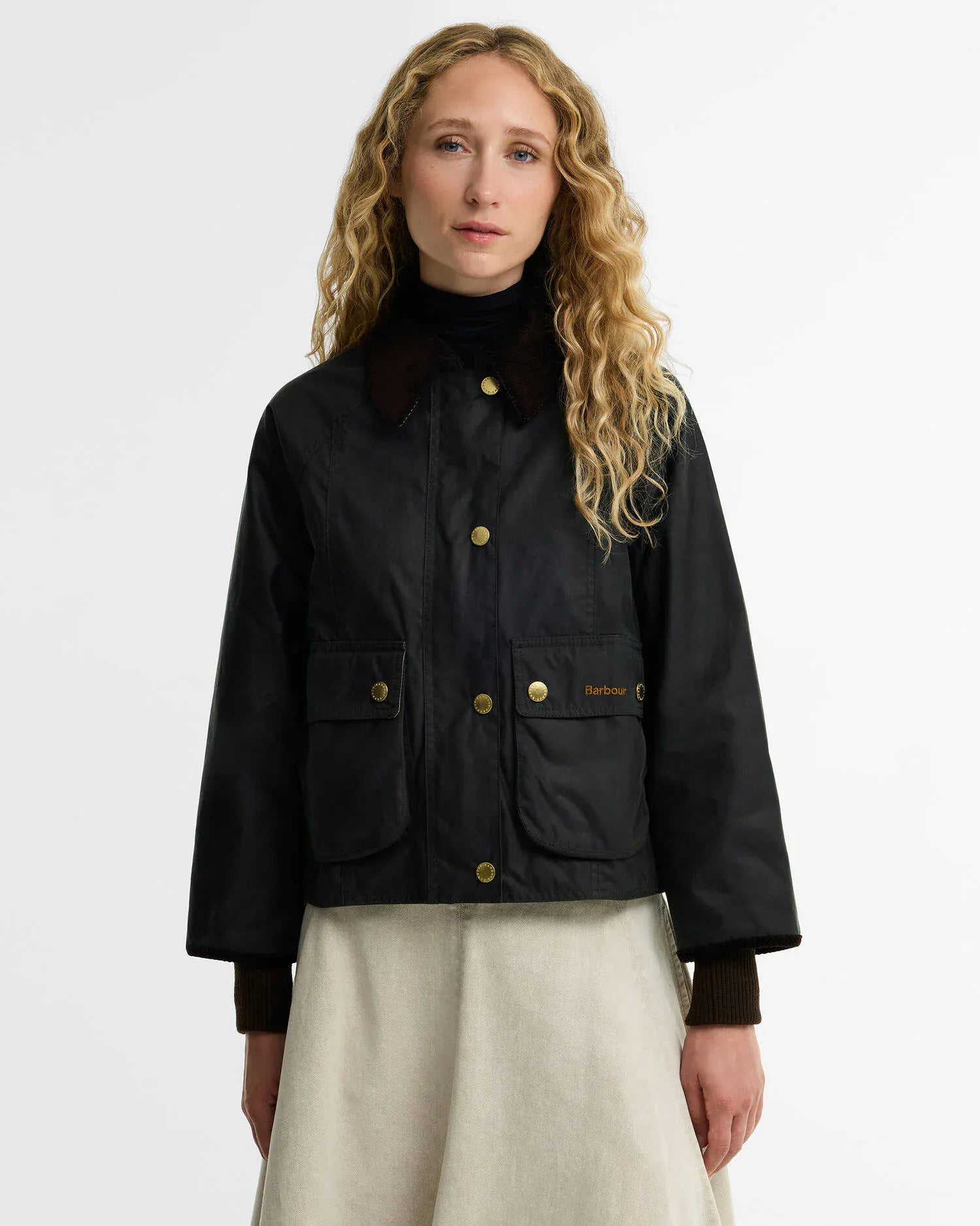 A model with long curly blonde hair wearing a black waxed cotton Barbour jacket with gold buttons and a dark brown corduroy collar, paired with a light beige skirt, against a plain white studio background.