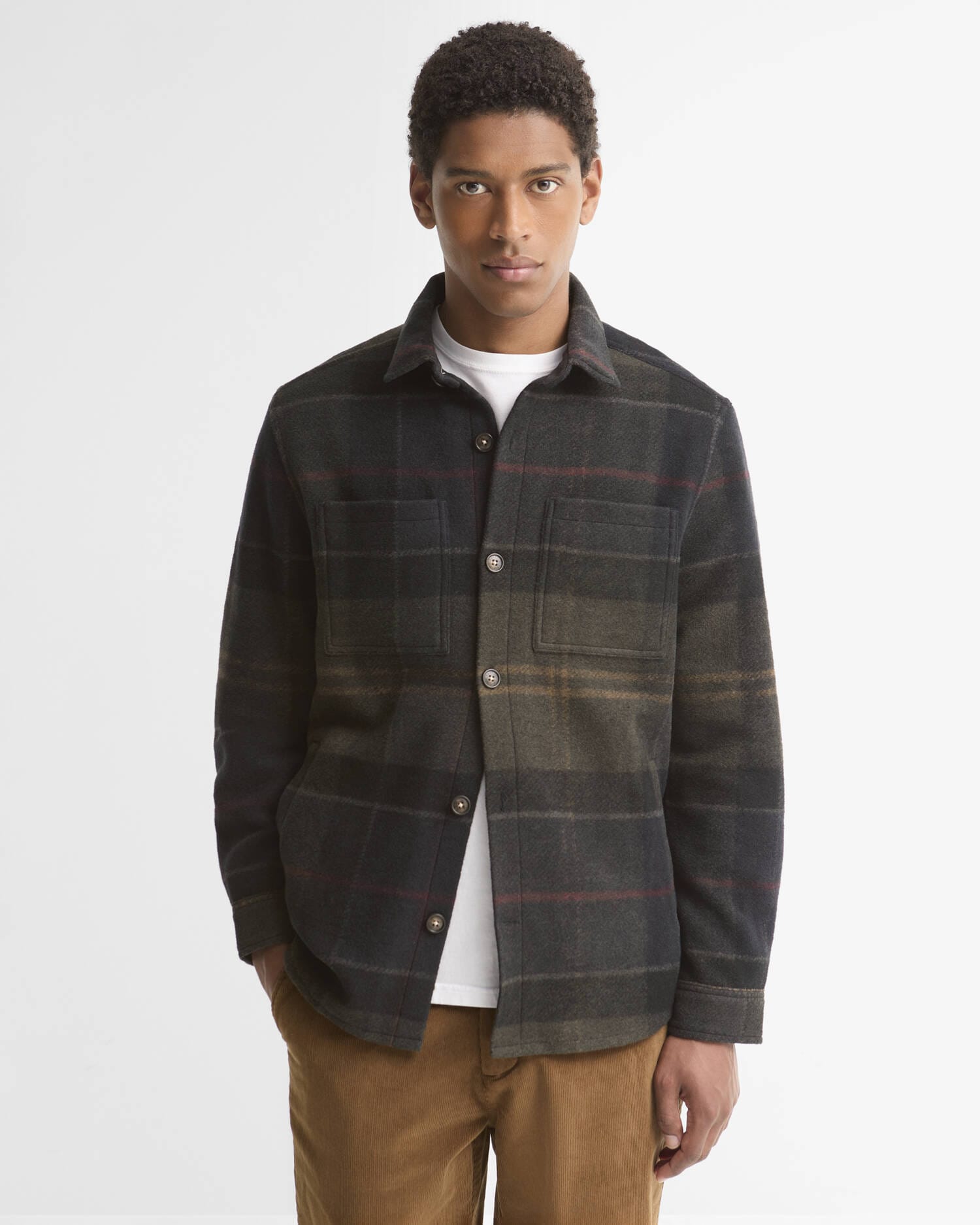 A man wearing a dark plaid button-up overshirt with brown and red stripes, layered over a white t-shirt and brown pants, standing against a light gray studio background.