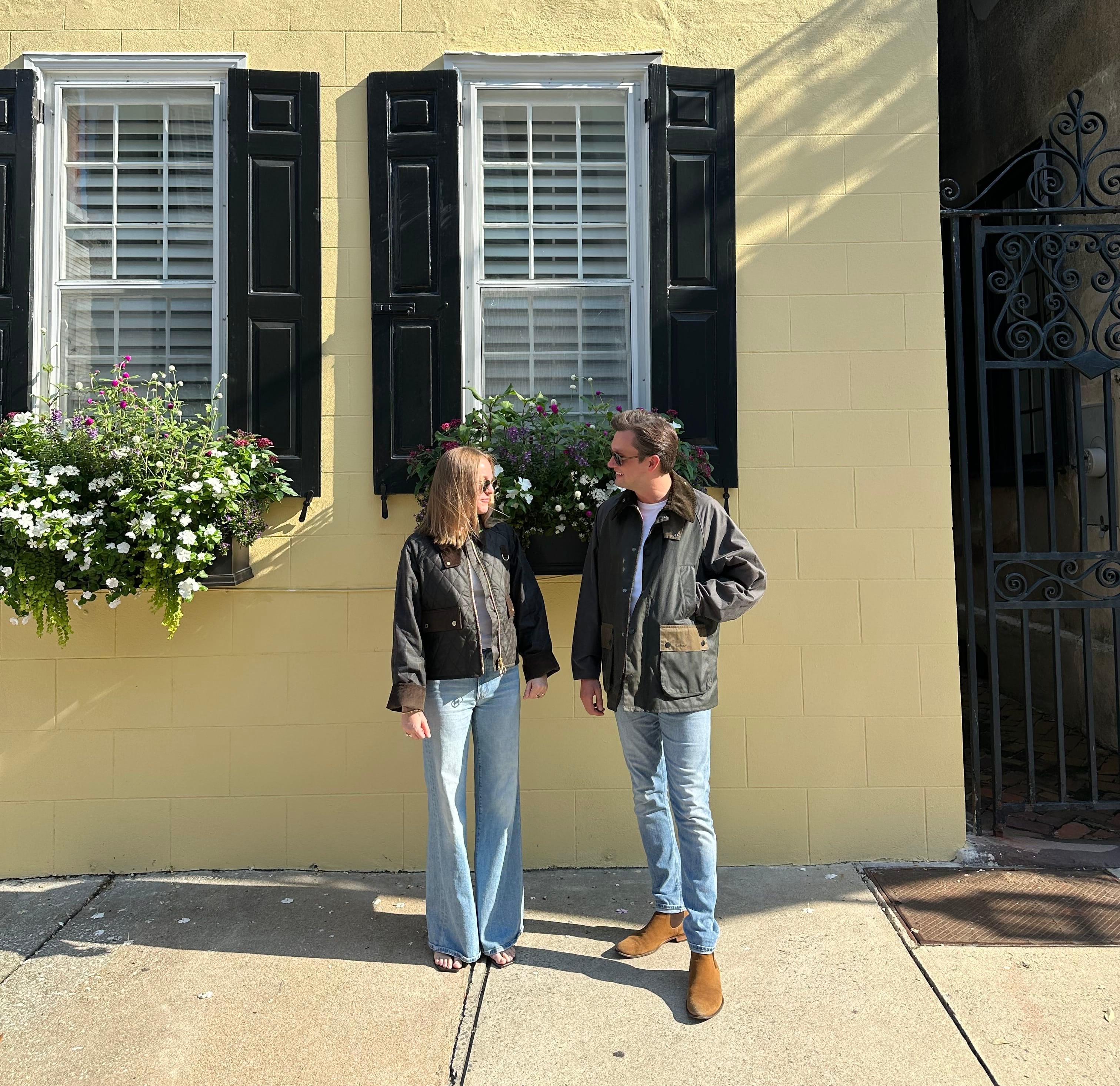 A man and a woman standing on a sidewalk in front of a yellow house with black shutters and flower boxes. The woman wears a black jacket and light blue wide-leg jeans, and the man wears a brown jacket, blue jeans, and brown boots.
