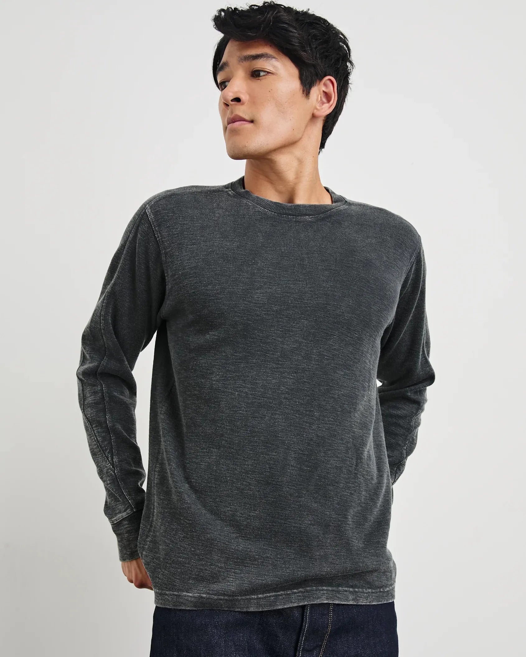 A man wearing a dark gray long-sleeve crewneck shirt and dark blue jeans, standing against a light gray studio background.
