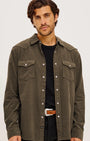 A man wearing an olive green corduroy button-up shirt jacket over a black t-shirt and black pants, standing against a light gray studio background.