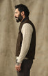 A man in profile wearing a dark brown sleeveless vest over a cream ribbed knit sweater, paired with dark trousers, standing against a textured beige backdrop.