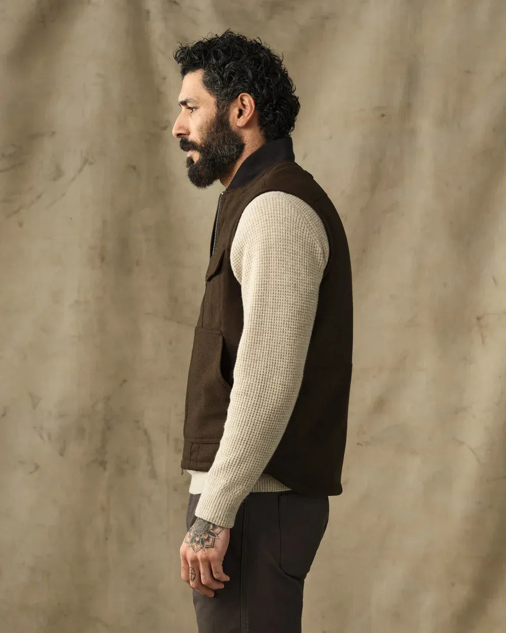 A man in profile wearing a dark brown sleeveless vest over a cream ribbed knit sweater, paired with dark trousers, standing against a textured beige backdrop.
