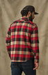 A man wearing a red, black, and beige plaid flannel shirt, dark blue jeans, and a dark brown baseball cap, shown from the back against a textured beige backdrop.
