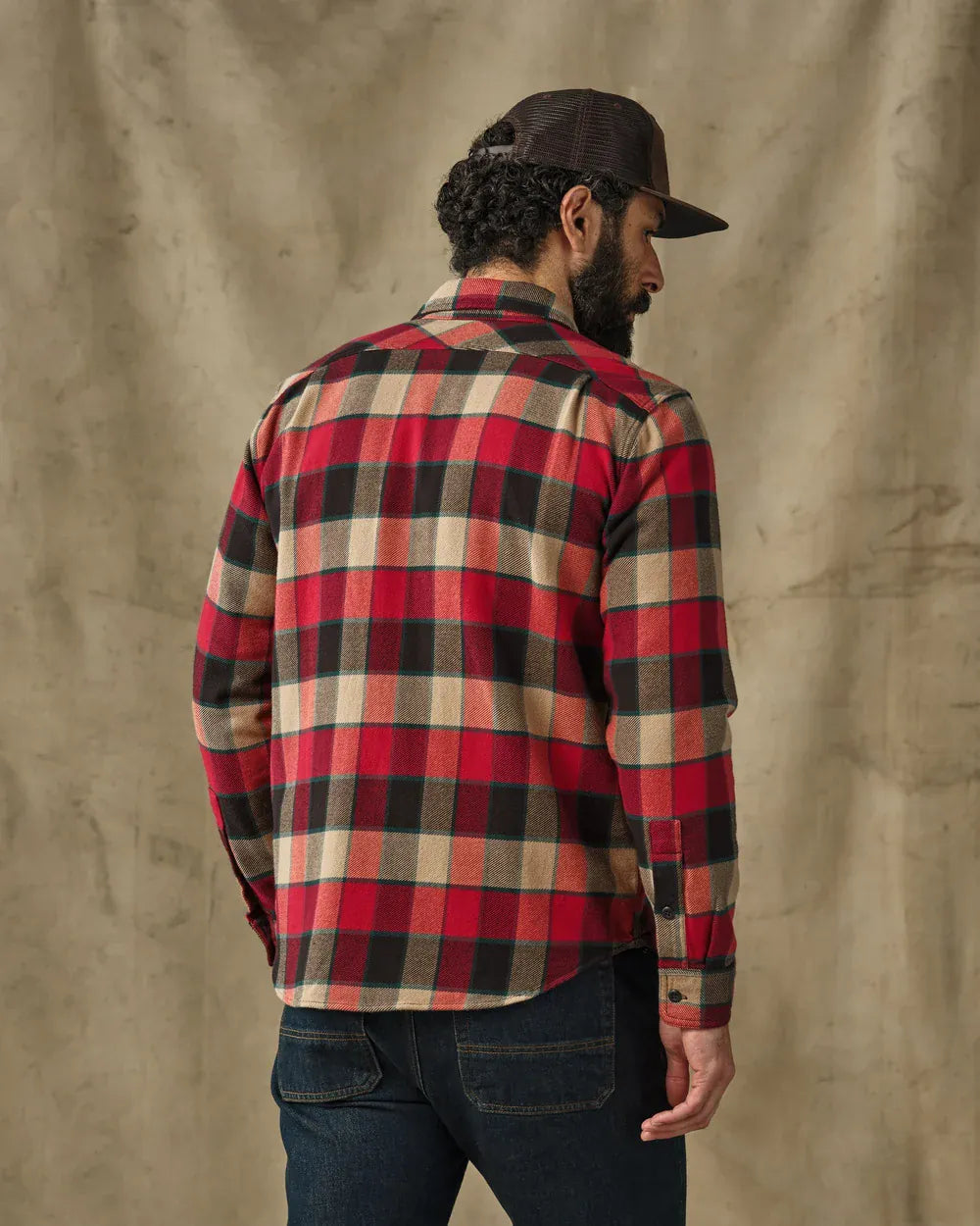 A man wearing a red, black, and beige plaid flannel shirt, dark blue jeans, and a dark brown baseball cap, shown from the back against a textured beige backdrop.