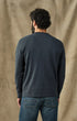 A man viewed from behind wearing a dark navy blue long-sleeve crewneck shirt and blue jeans, standing against a textured beige fabric backdrop.