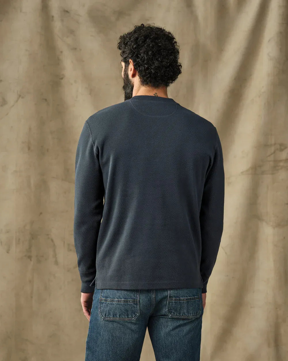 A man viewed from behind wearing a dark navy blue long-sleeve crewneck shirt and blue jeans, standing against a textured beige fabric backdrop.